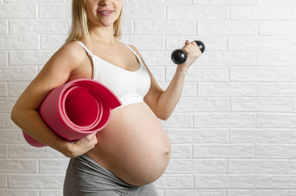 Pregnant woman holding a yoga mat and dumbbell, dressed in workout clothes, preparing for exercise